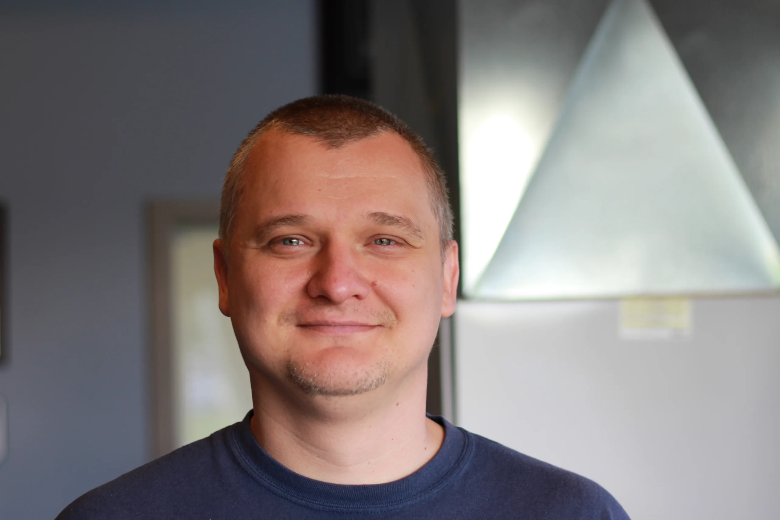 Dmytro Sereda, K Komfort Residential HVAC Installer A professional headshot of Dmytro Sereda, an EPA-certified residential HVAC installer at K Komfort, smiling with sheet metal ductwork in the background.