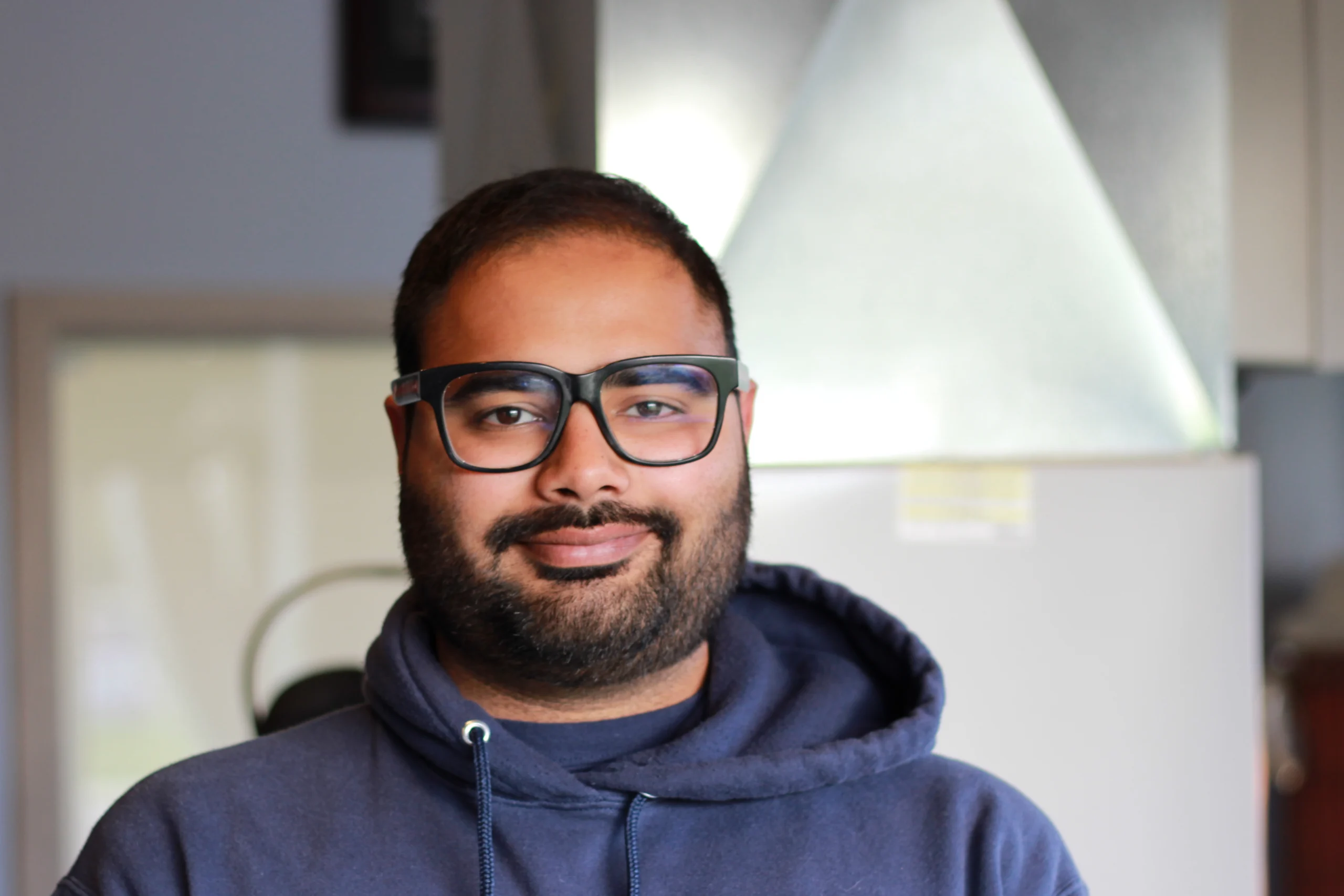 Gurman Singh, K Komfort HVAC Technician A professional headshot of Gurman Singh, an EPA-certified HVAC service technician at K Komfort, wearing thick black glasses and a blue hoodie with indoor sheet metal ductwork in the background.