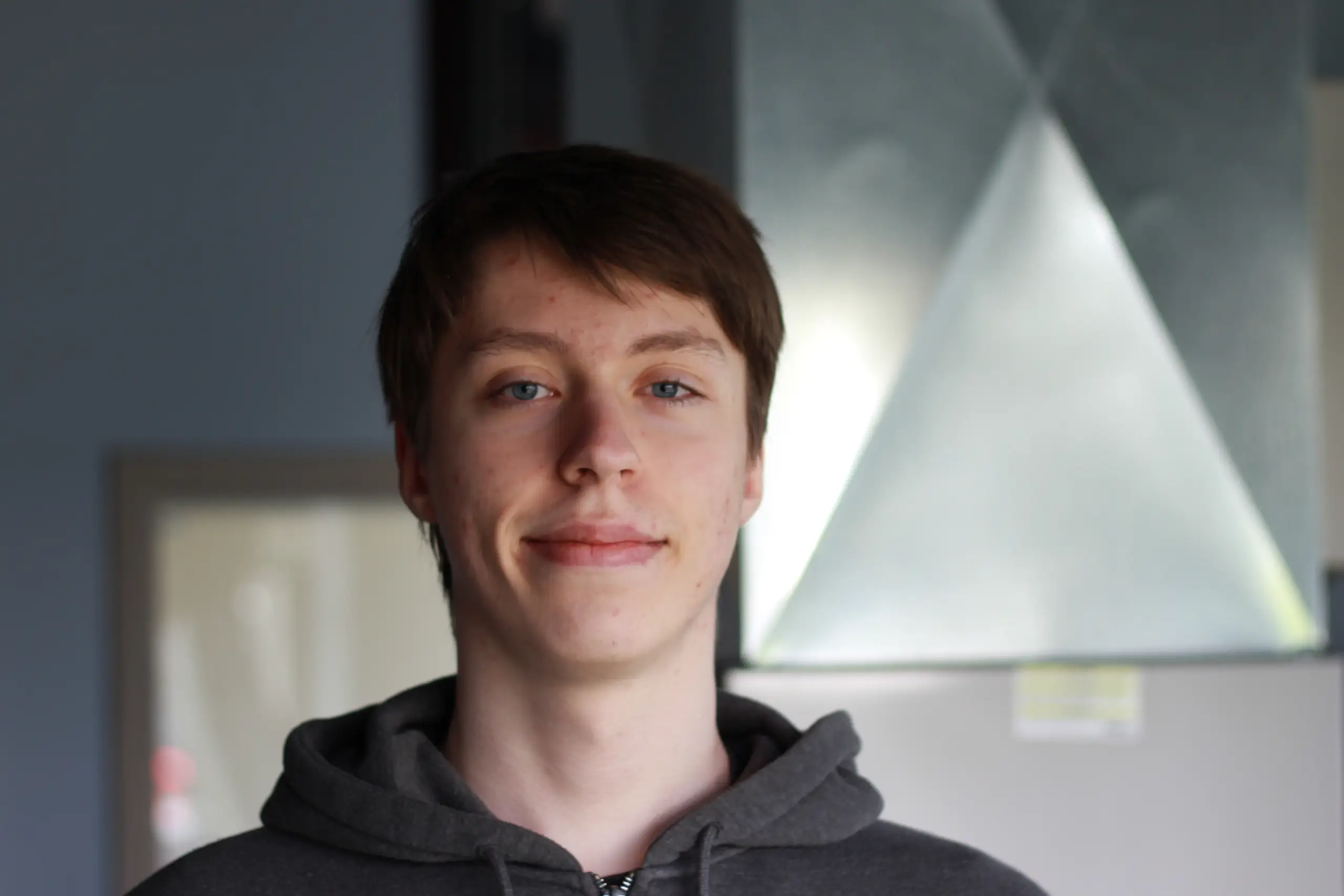 Headshot of Ilya Prihod, an Assistant HVAC Designer at K Komfort, smiling in an office or training environment with ventilation equipment visible in the background.