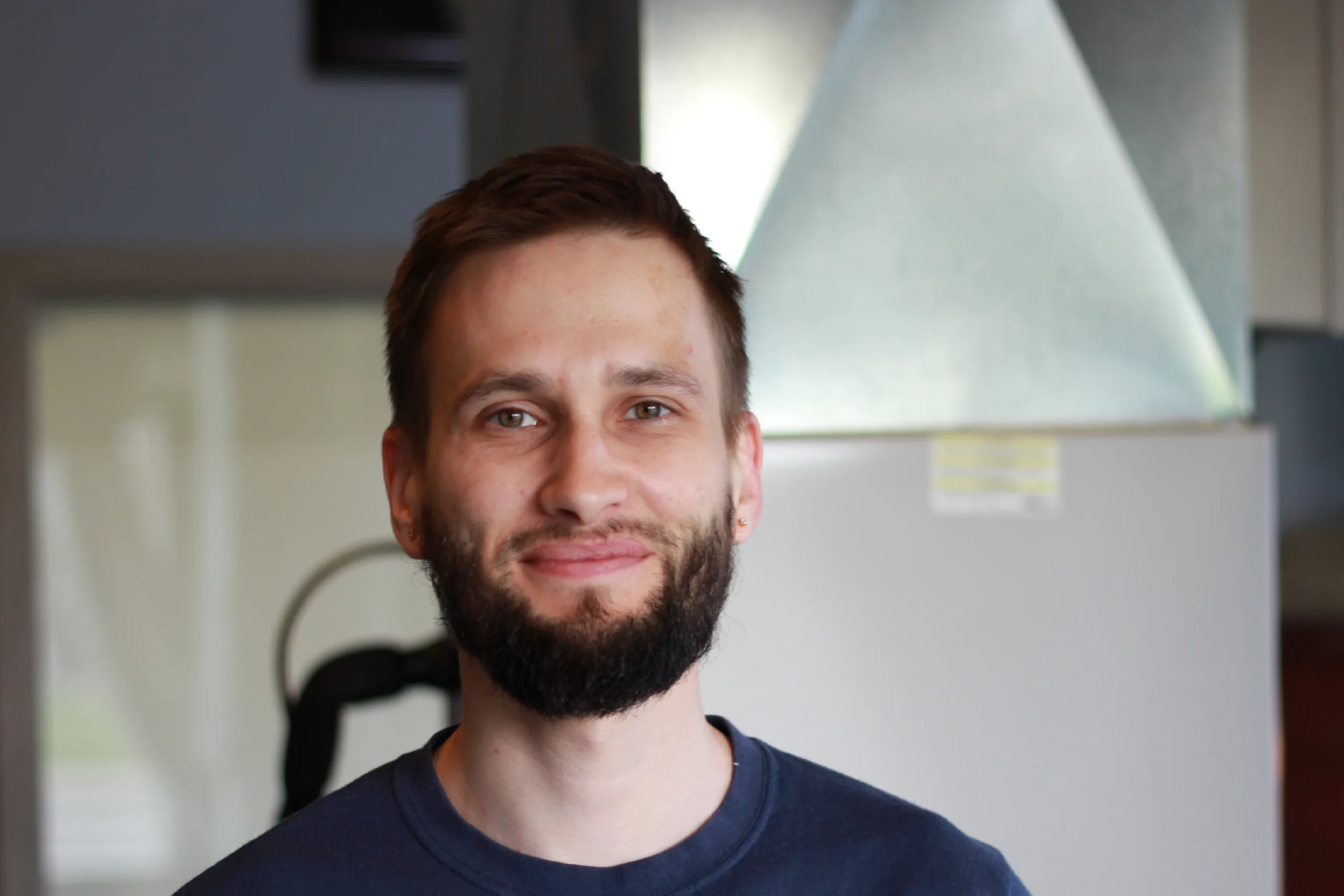 Maksym Liakhov, Residential HVAC Installation Lead A professional headshot of Maksym Liakhov, an EPA-certified residential HVAC installation crew lead at K Komfort, smiling in front of sheet metal ductwork.