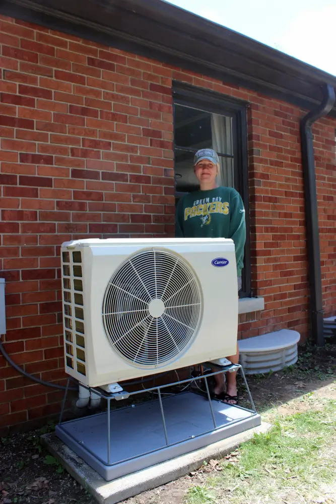 Carrier Heat Pump Upgrade Installation A smiling homeowner in a Green Bay Packers sweatshirt standing behind a newly installed, slim side-discharge Carrier heat pump system mounted on an elevated winter snow stand outside a brick home in Milwaukee, Wisconsin.