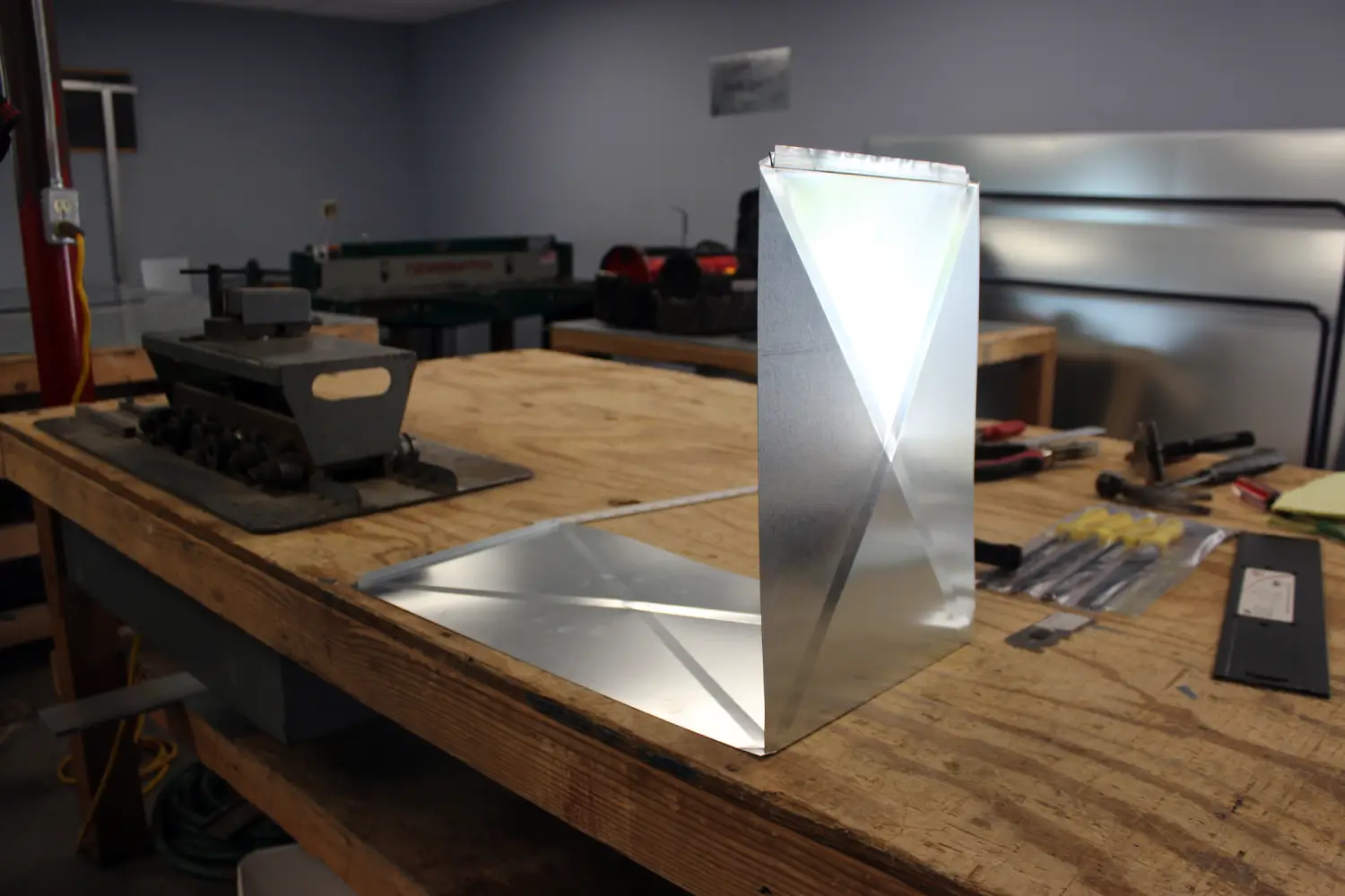 Custom-bent galvanized sheet metal ductwork component on a workshop wooden bench with hand tools at K Komfort in Grafton WI. Custom-bent galvanized sheet metal ductwork component on a workshop wooden bench with hand tools at K Komfort in Grafton WI.