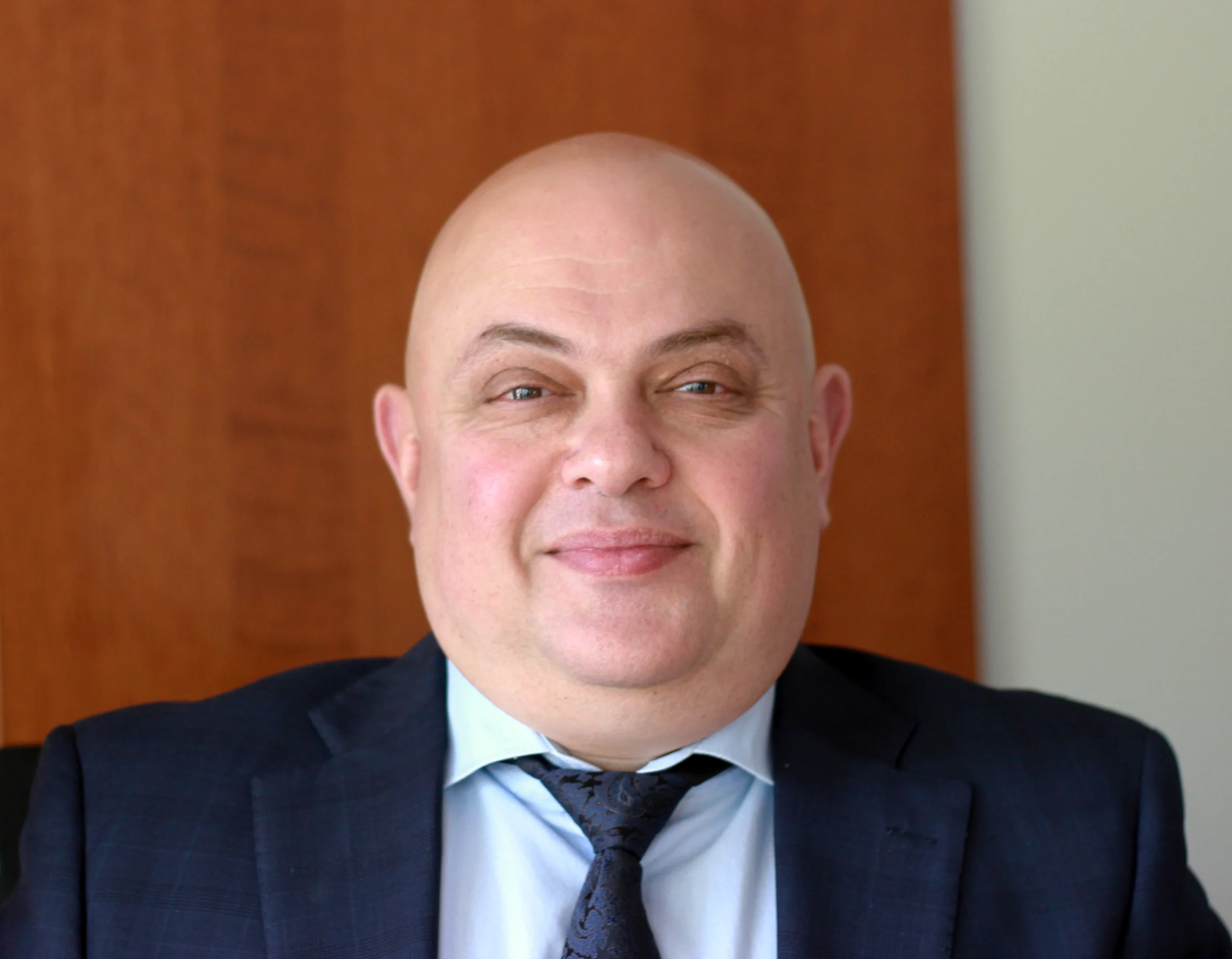 Eugene Krol, K Komfort General Manager A professional portrait of Eugene Krol, General Manager of K Komfort Heating & Cooling, wearing a dark suit jacket and tie against a warm wood-paneled background.