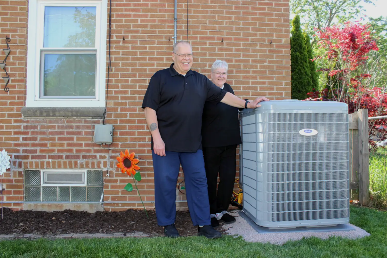 Satisfied HVAC Upgrade Customers Smiling Wisconsin homeowners standing outside their brick home next to a newly installed Carrier heat pump system upgraded through the HEAR rebate program by K Komfort in Milwaukee.