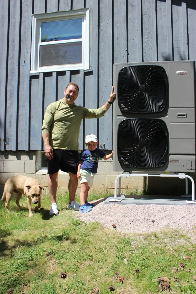 Heat Pump Upgrade in Grafton, WI A smiling local homeowner and his young son standing with their dog next to a newly installed side-discharge Lennox heat pump system mounted on an elevated ground stand outside a house in Grafton, Wisconsin.