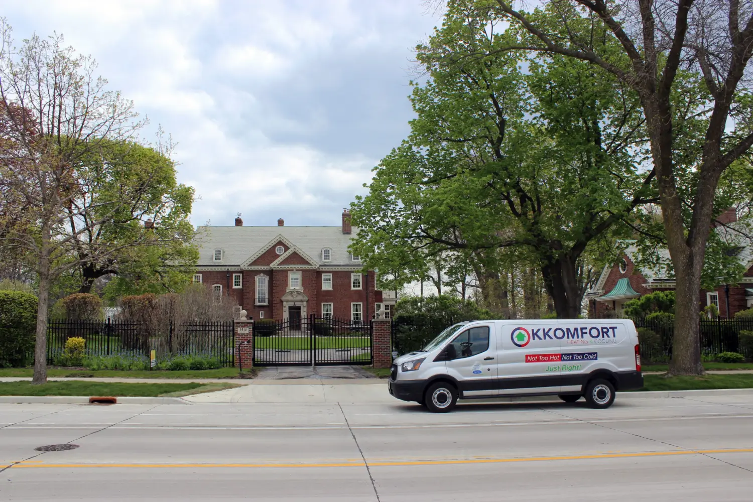 K Komfort Historic Colonial HVAC Service in Whitefish Bay A silver K Komfort Heating & Cooling service van driving past a large historic red brick Georgian Colonial mansion with black wrought-iron fencing in Whitefish Bay, Wisconsin.
