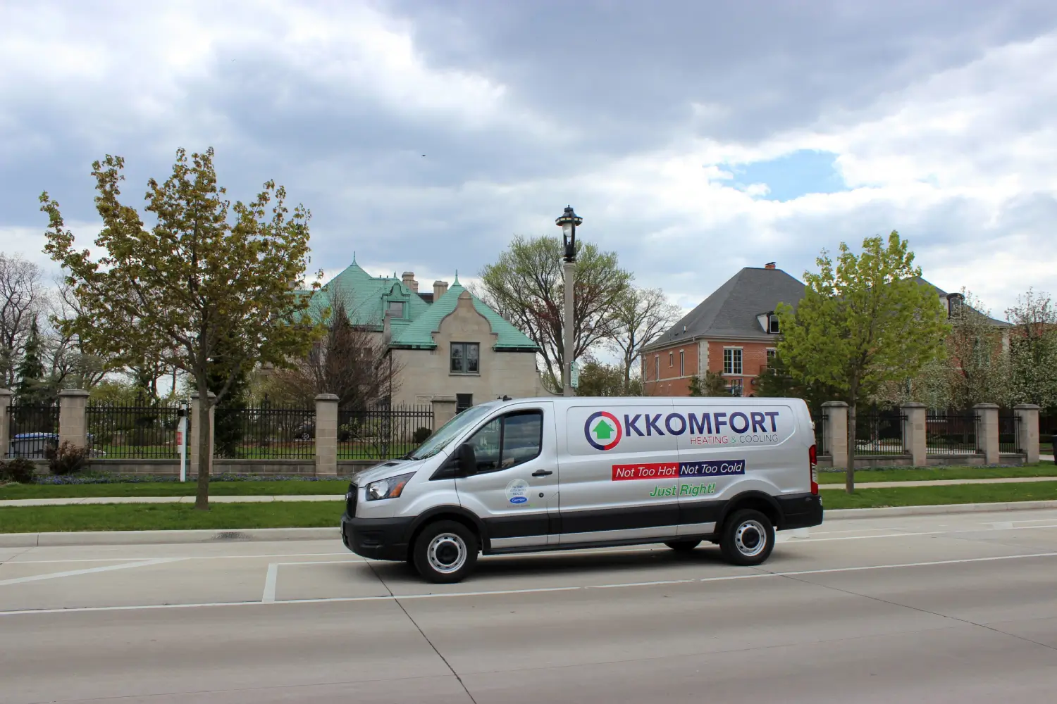 K Komfort Historic Estate HVAC Service in Whitefish Bay A silver K Komfort Heating & Cooling service van driving on a street past a historic stone mansion with a green copper-clad roof and stone pillar fencing in Whitefish Bay, Wisconsin.
