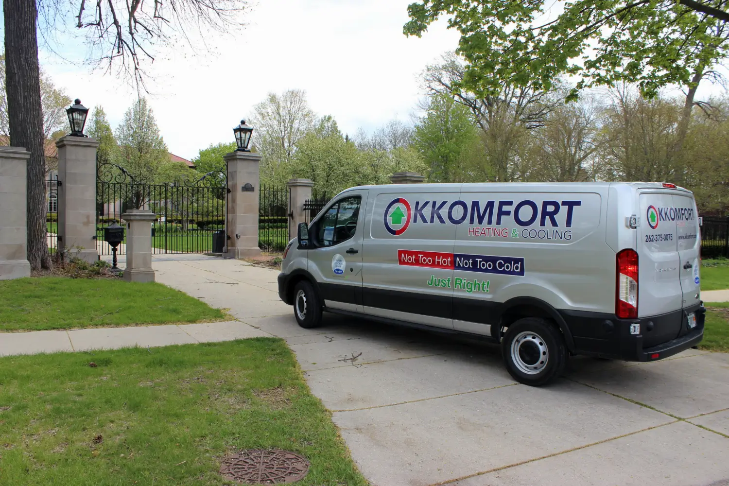 K Komfort Historic Home HVAC Service in Whitefish Bay A silver K Komfort Heating & Cooling service van parked in the driveway of a historic North Shore estate with grand stone pillars and wrought-iron gates in Whitefish Bay, Wisconsin.