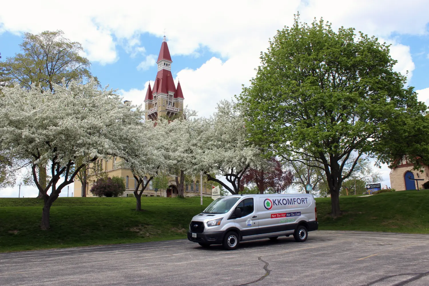 K Komfort HVAC Van in West Bend near the Tower Heritage Center A silver K Komfort Heating & Cooling service van parked in West Bend, Wisconsin, with the historic red tower of the Tower Heritage Center and Fifth Avenue United Methodist Church visible behind spring trees.