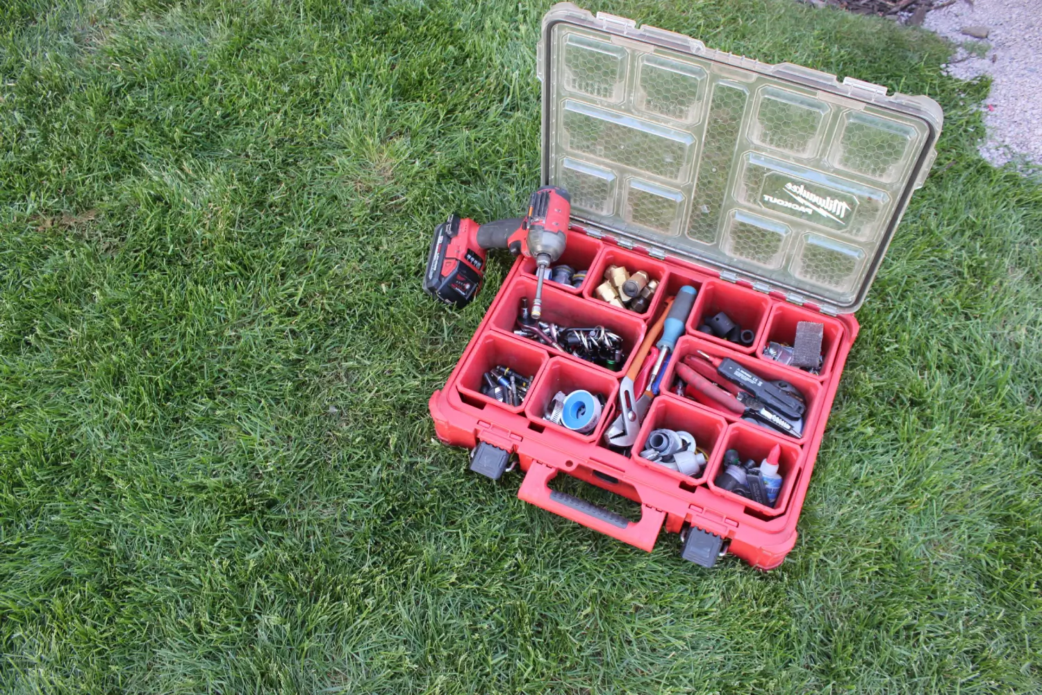 HVAC Maintenance Tool Setup An open Milwaukee Tool PACKOUT organizer box containing HVAC technician tools, brass plumbing fittings, and electrical components sitting on green grass during a preventative maintenance plan visit by K Komfort in Milwaukee, Wisconsin.