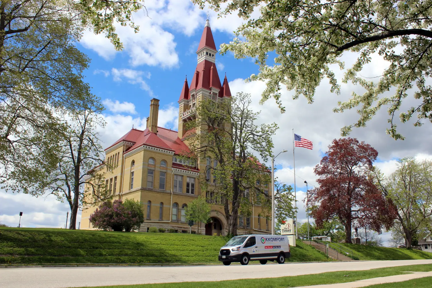 A silver K Komfort Heating & Cooling service van driving past the historic Old Washington County Courthouse tower, now the Tower Heritage Center, on a sunny day in West Bend, Wisconsin.