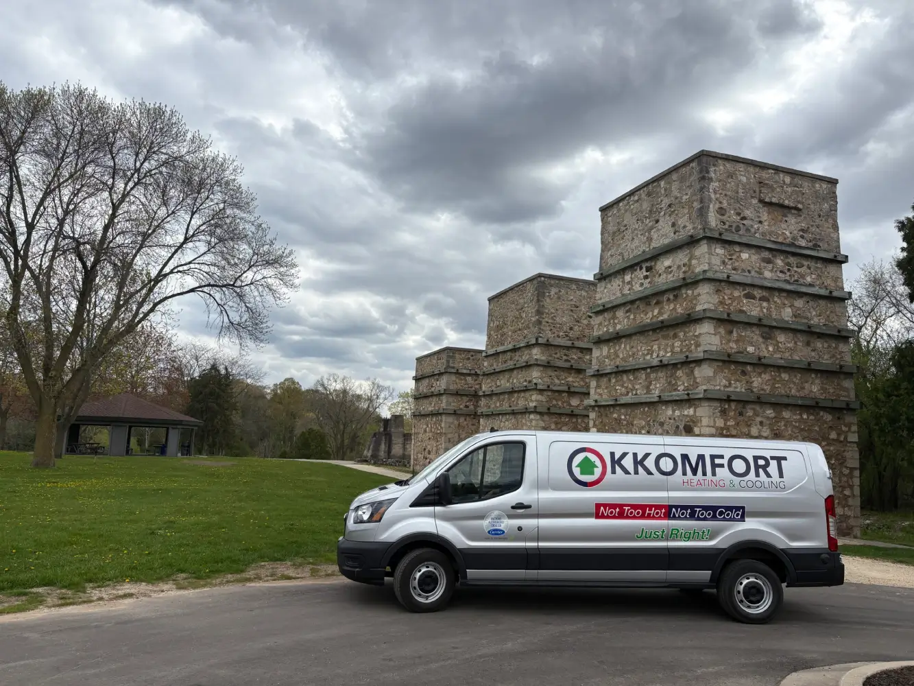 A silver K Komfort Heating & Cooling service van parked along the road at Lime Kiln Park in Grafton, Wisconsin, with the historic stone lime kiln structures and park pavilion in the background under a cloudy sky.