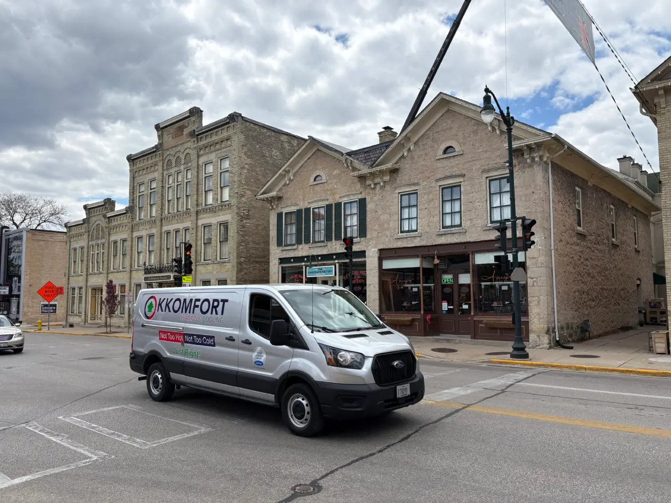 K Komfort HVAC Service in Cedarburg, WI A silver K Komfort Heating & Cooling service van driving through the historic intersection of Washington Avenue in downtown Cedarburg, Wisconsin, with the Washington House Inn and local brick storefronts in the background.