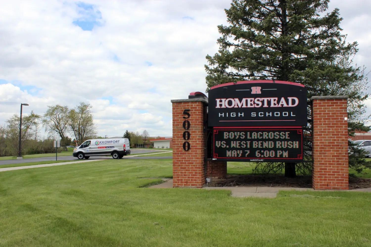 K Komfort HVAC Service in Mequon, WI A silver K Komfort Heating & Cooling service van driving past the brick entrance sign of Homestead High School on Mequon Road in Mequon, Wisconsin.