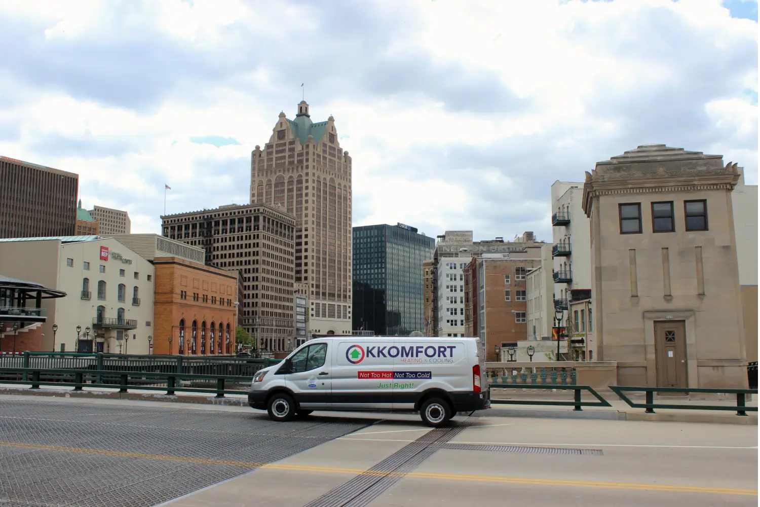 Local HVAC Service Van in Downtown Milwaukee A white K Komfort Heating & Cooling service van driving across a bridge over the river with the downtown Milwaukee skyline and historic brick buildings in the background.