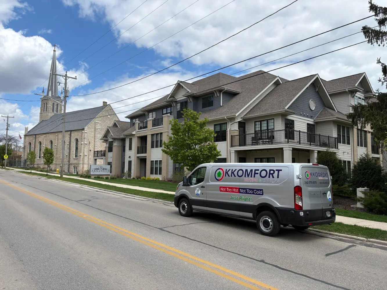 K Komfort HVAC Service on Washington Ave in Cedarburg A silver K Komfort Heating & Cooling service van parked along Washington Avenue in Cedarburg, Wisconsin, with the Arrabelle apartments and the historic stone steeple of First Immanuel Lutheran Church in the background.