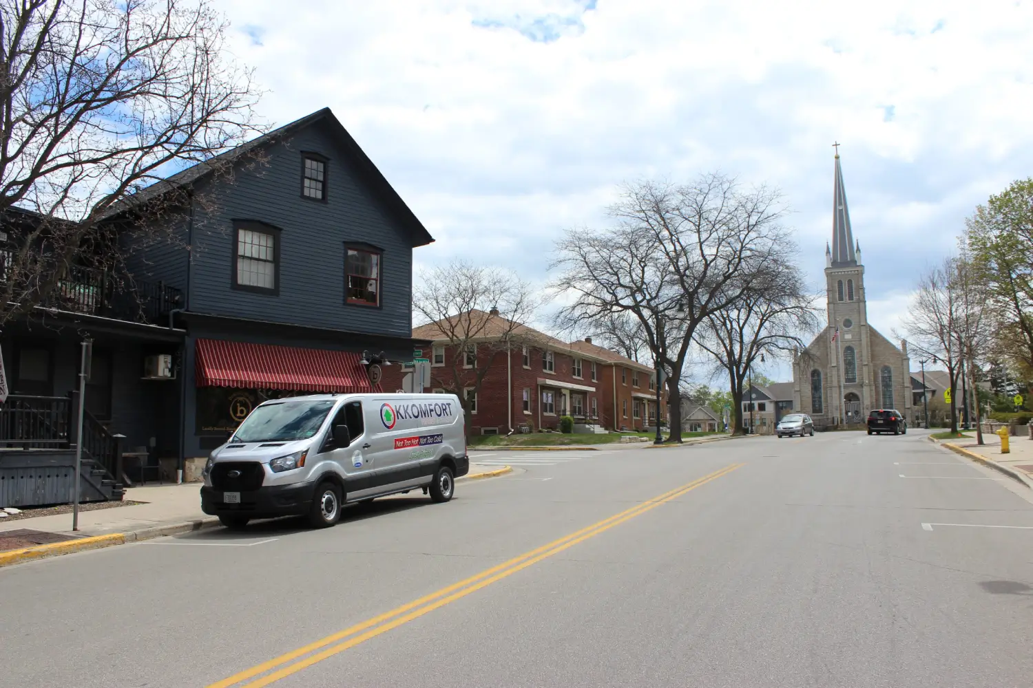 K Komfort HVAC Service on Washington Avenue A silver K Komfort Heating & Cooling service van parked along Washington Avenue in Cedarburg, Wisconsin, with local residential buildings and the historic stone steeple of First Immanuel Lutheran Church visible in the background.