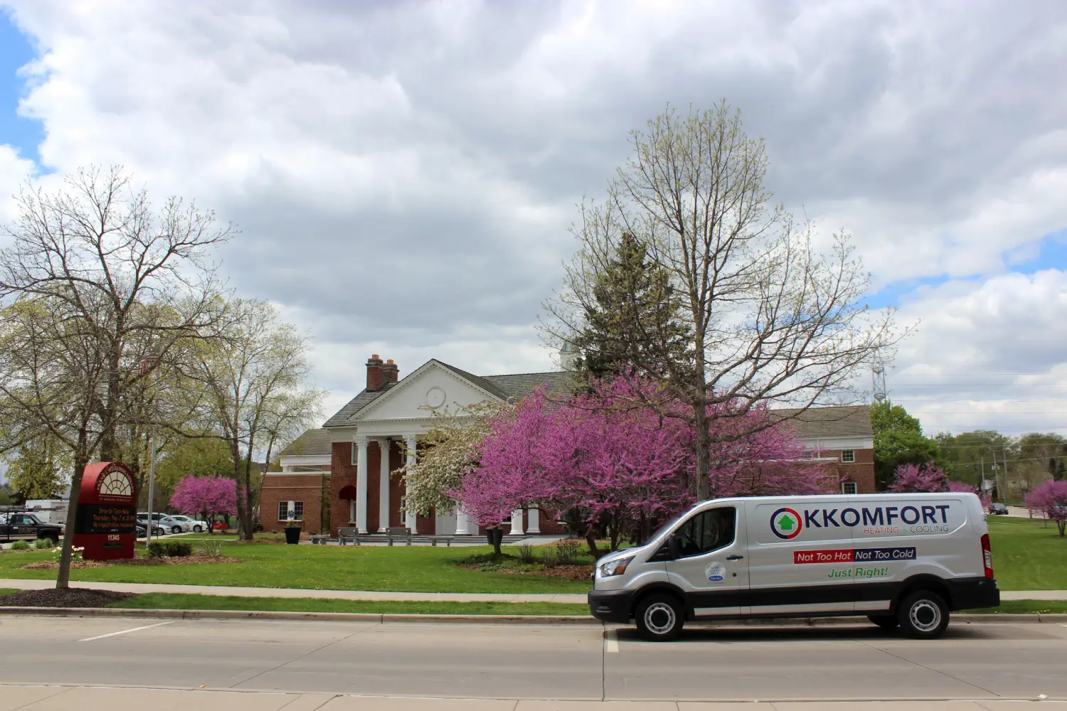 K Komfort HVAC Van at Frank L Weyenberg Library A silver K Komfort Heating & Cooling service van parked along the street in front of the Frank L. Weyenberg Library building framed by blooming pink trees in Mequon, Wisconsin.
