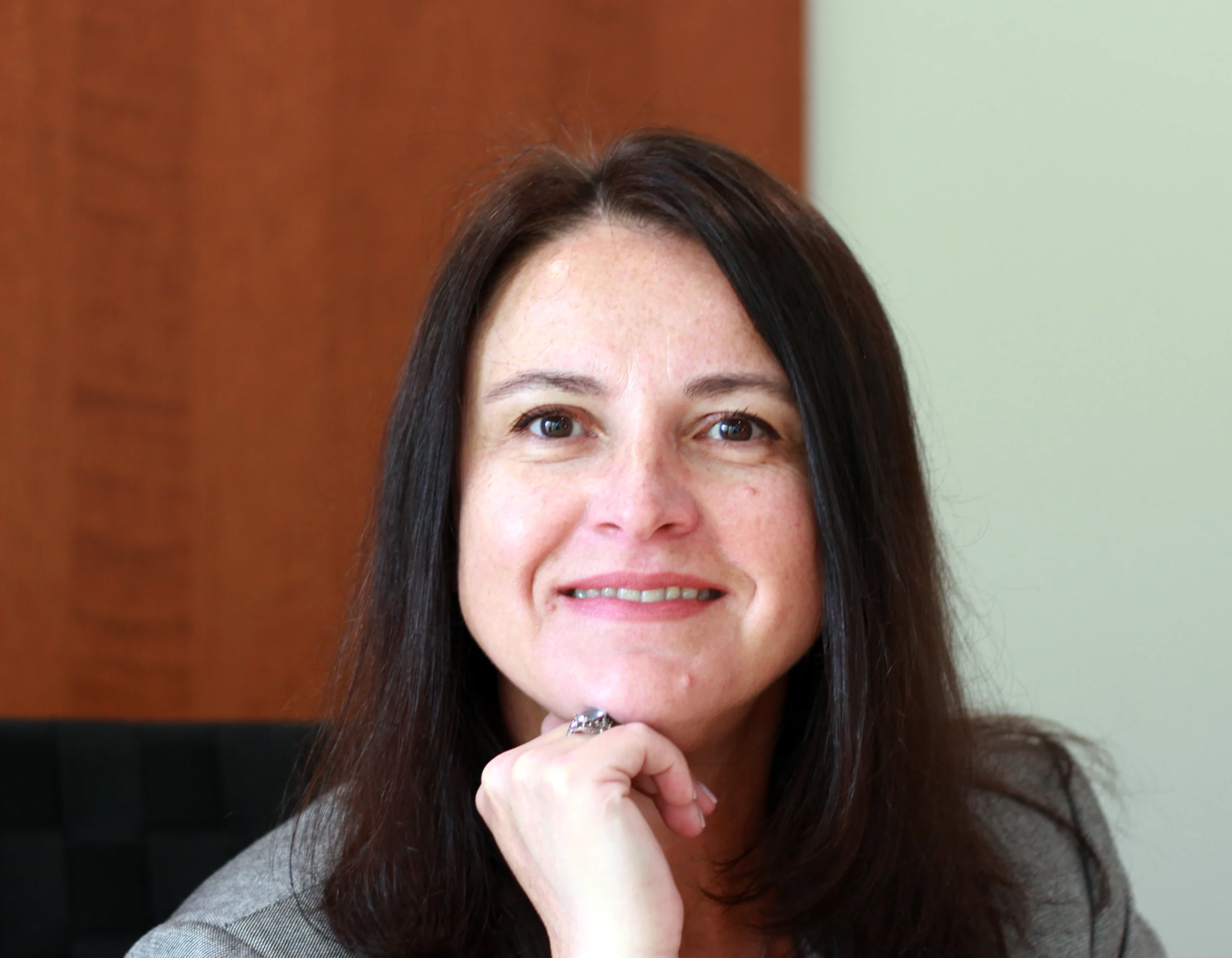 Irina Krol, K Komfort CFO A professional portrait of Irina Krol, CFO of K Komfort Heating & Cooling, smiling with her hand resting under her chin against a warm wood-paneled background.
