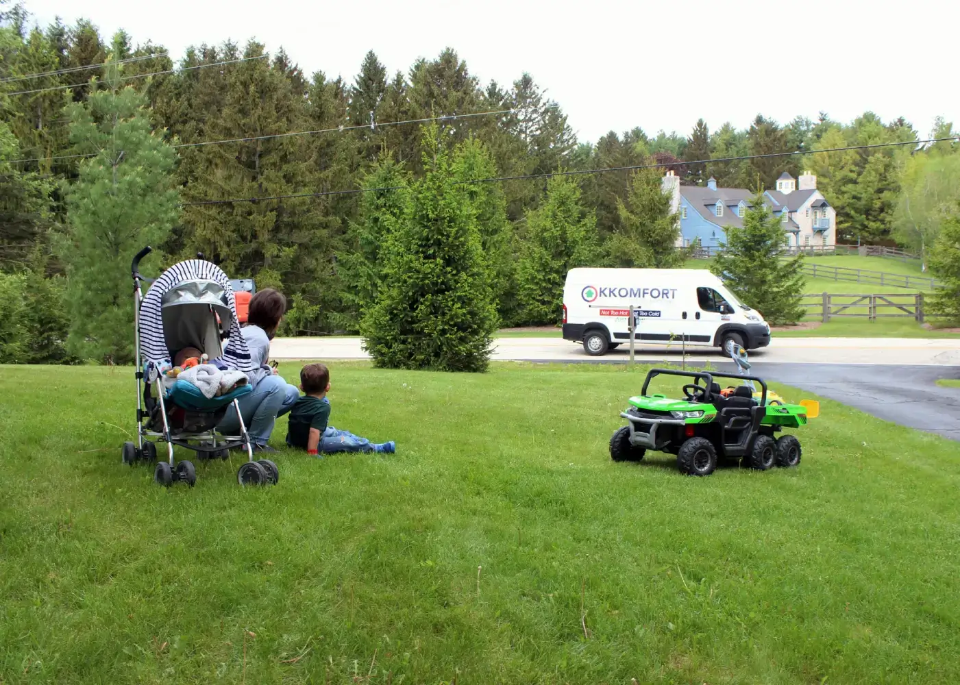 Trusted Neighborhood HVAC Services A mother and young child sitting on a green residential lawn next to a baby stroller, watching a white K Komfort Heating & Cooling service van drive down a neighborhood street in Grafton, Wisconsin.