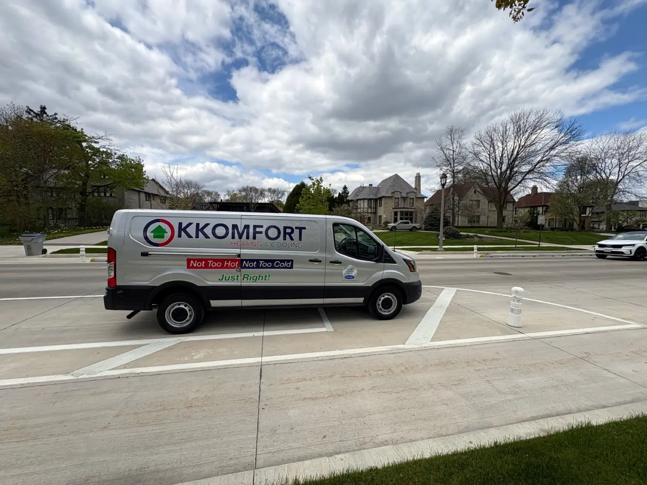 K Komfort Historic Tudor HVAC Service in Whitefish Bay A silver K Komfort Heating & Cooling service van parked along a white-lined street median across from historic stone Tudor Revival homes in Whitefish Bay, Wisconsin.
