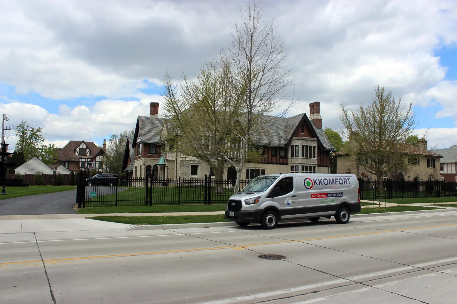 K Komfort Historic Tudor HVAC Service in Whitefish Bay A silver K Komfort Heating & Cooling service van parked on the street in front of a historic stone and half-timbered Tudor Revival mansion in Whitefish Bay, Wisconsin.