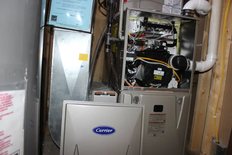 A K Komfort technician inspecting the internal components of an open Carrier high-efficiency gas furnace during a tune-up service.