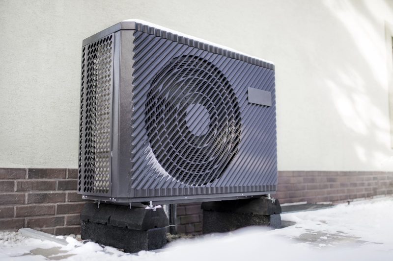 How to Determine Heat Pump Efficiency - Photo of a modern air source heat pump unit standing against the residential building wall. Shot in winter, in a snowy setting.