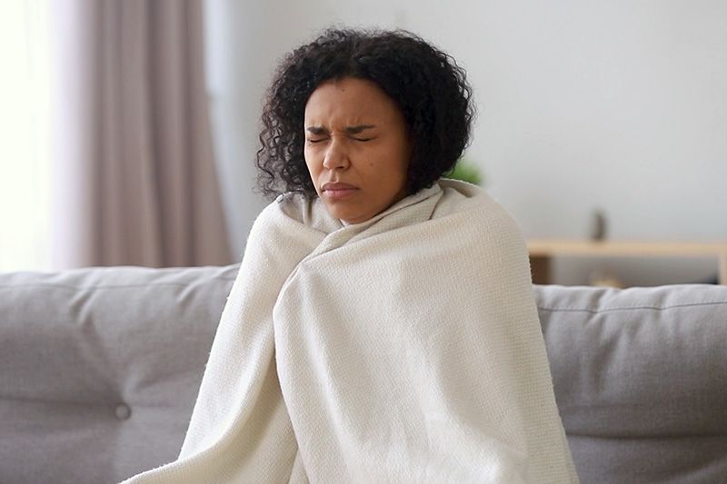 Video: Upgrade Your Furnace. Image shows woman sitting on a couch wrapped in a blanket.